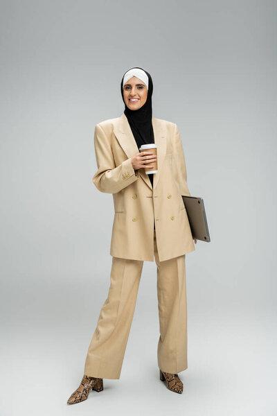 joyful muslim businesswoman in hijab and beige suit standing with takeaway drink and laptop on grey