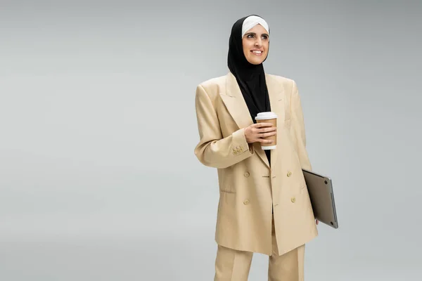 stock image delighted and fashionable muslim businesswoman with paper cup and laptop looking away on grey