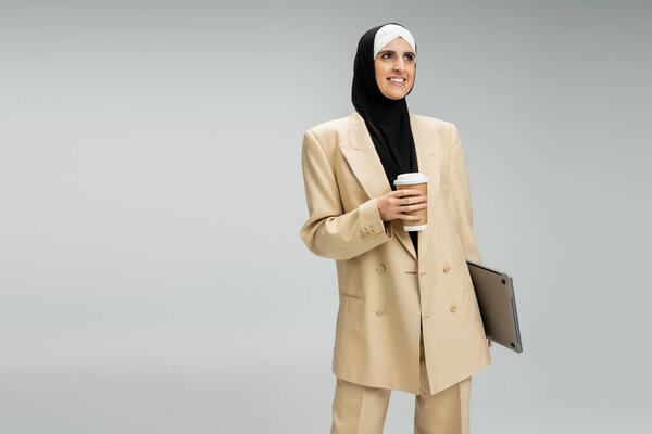 delighted and fashionable muslim businesswoman with paper cup and laptop looking away on grey