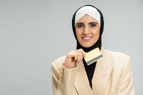 stylish muslim businesswoman in blazer and hijab holding credit card and smiling on grey, headshot