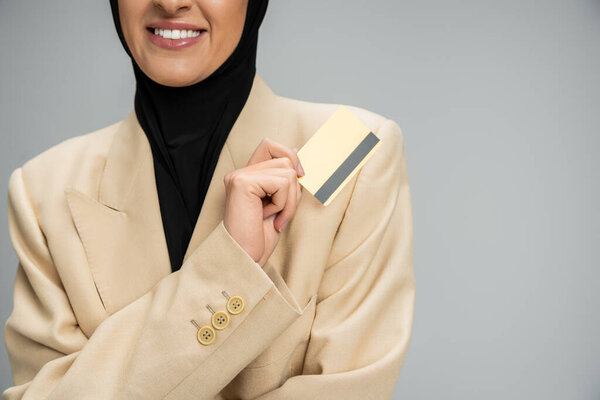 cropped view of smiling muslim businesswoman in blazer and hijab holding credit card on grey