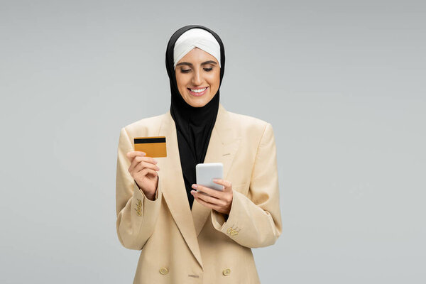 pleased muslim businesswoman in hijab and stylish blazer holding smartphone and credit card on grey