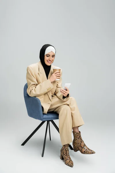 stock image joyful and stylish muslim businesswoman sitting in armchair with paper cup and smartphone on grey