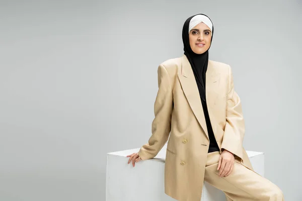 stock image successful muslim businesswoman in stylish suit and hijab smiling at camera near white cube on grey