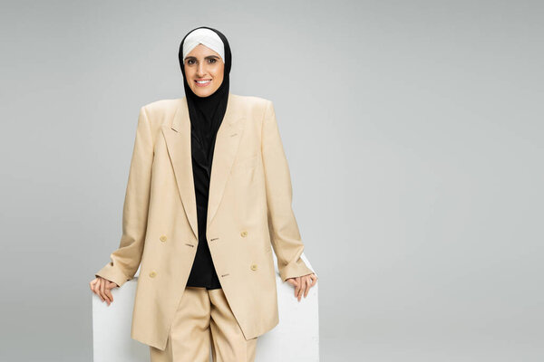 happy muslim businesswoman in beige suit and hijab smiling at camera near white cube on grey