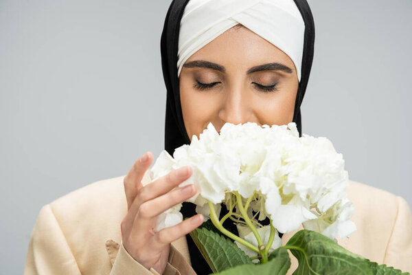 stylish and modern muslim businesswoman with closed eyes smelling hydrangea flower on grey