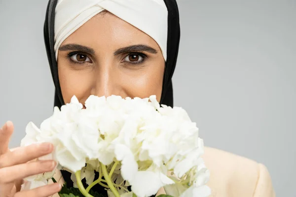delighted muslim businesswoman obscuring face with hydrangea flower and looking at camera on grey