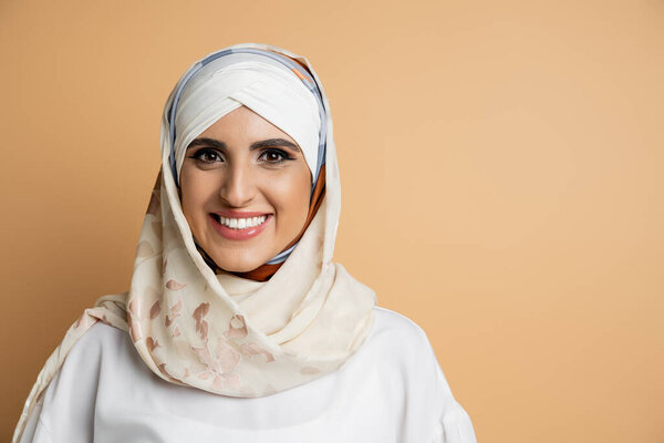 smiling muslim woman with makeup wearing elegant headscarf and looking at camera on beige, portrait