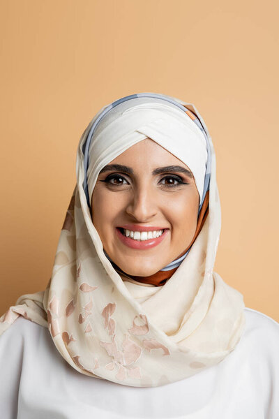 portrait of charming muslim woman with makeup, in silk headscarf smiling at camera on beige