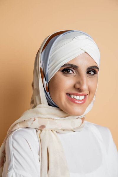 joyful muslim woman in elegant silk headscarf looking at camera on beige, portrait with makeup