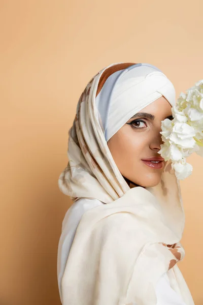 charming muslim woman in headscarf obscuring face with white flower and looking at camera on beige