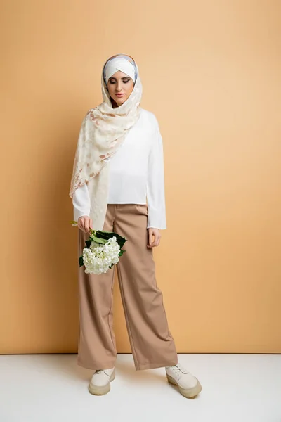 stock image modern muslim woman in silk headscarf and stylish casual attire posing with white flower on beige