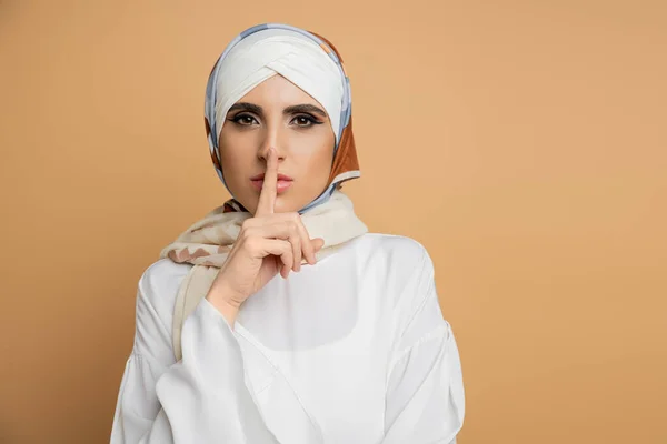 stock image fashionable muslim woman in silk headscarf showing hush gesture and looking at camera on beige