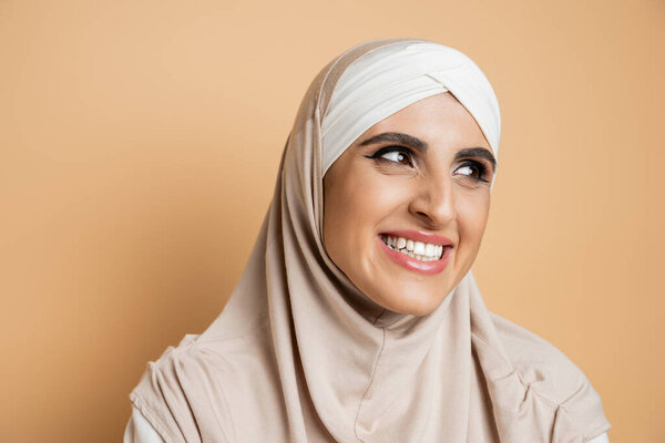 cheerful and elegant muslim woman with makeup wearing hijab and looking away on beige