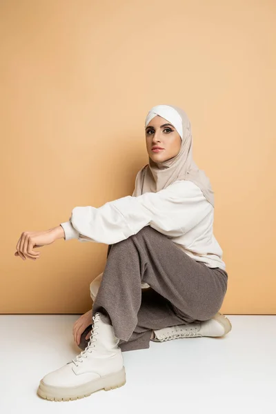 stock image confident muslim woman in hijab, grey pants and leather boots sitting and looking at camera on beige