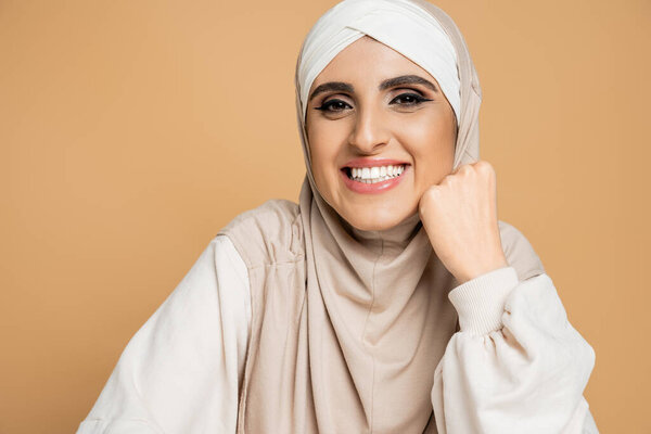 happy muslim woman in hijab and white sweatshirt smiling at camera on beige, modern lifestyle