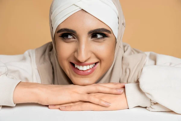 portrait of cheerful muslim woman in hijab sitting at white table and looking away on beige