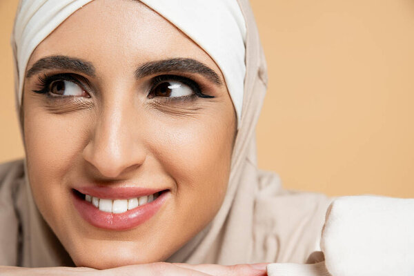close up portrait of smiling muslim woman with makeup, in hijab, looking away on beige