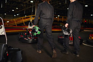 Yarış arabalarının yanında yürüyen ve ellerinde miğfer tutan iki yarışçı, go-kart sürücüleri.