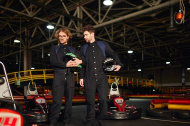 two kart racers standing near racing cars and holding helmets, male drivers in kart circuit clipart