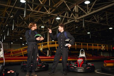 kart racer pointing at friend in glasses and standing near racing cars while holding helmets clipart