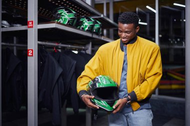 Sarı bombardıman ceketli, kasklı, erkek sürücü, go-cart 'lı mutlu Afro-Amerikan araba yarışçısı.