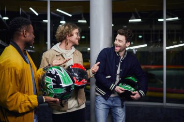 cheerful interracial male friends in jackets holding helmets, indoor racing track, karting concept clipart