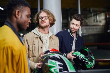 men with helmets looking at african american friend inside of indoor racing track, karting clipart