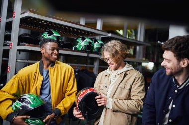 joyful interracial male friends in jackets holding helmets, indoor racing track, karting concept clipart