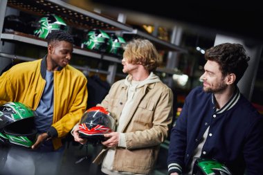 happy multicultural male friends in jackets holding helmets, indoor racing track, karting concept clipart