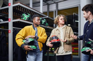 happy multicultural male friends holding helmets and talking inside of indoor racing track, karting clipart