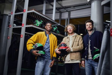 happy multicultural male friends holding helmets and looking at camera inside of indoor racing track clipart