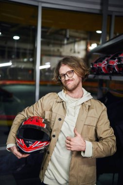 Gözlüklü, miğferli mutlu bir adam. Kapalı karting pistinde başparmağını gösteriyor. Hobi olarak.