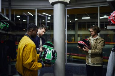 three multicultural friends in casual clothes choosing helmets for karting, go-cart concept clipart