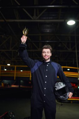 Spor giyimli neşeli bir sürücü elinde altın kupayla go-kart yarışının galibi olarak ayakta duruyor.