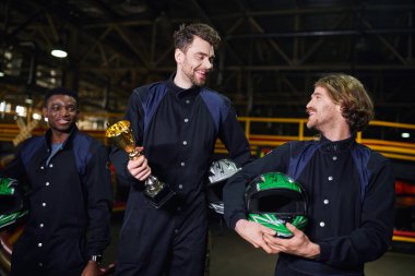 excited man holding golden cup and standing as a winner of go-kart race near diverse drivers clipart