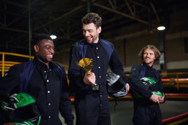 Altın kupa tutan neşeli bir adam ve farklı sürücülere yakın go-kart yarışının galibi olarak ayakta duruyor.