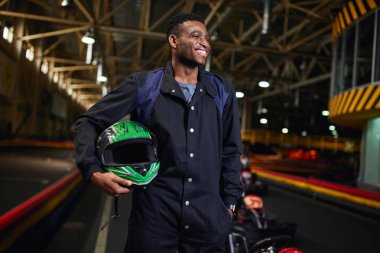 Spor kıyafetli neşeli Afrikalı Amerikalı adam kaskını tutuyor ve karting pistinde go-kart 'ın yanında duruyor.