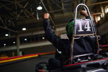 back view of motorsports driver in helmet raising hand and winning race on circuit, go-kart clipart