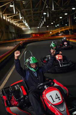 Heyecanlı Afro-Amerikan go cart sürücüsü kaskını takıyor, el kaldırıyor ve pistte yarışı kazanıyor, go-kart