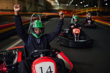 Heyecanlı Afro-Amerikan go kart yarışçısı kaskını takıp el kaldırıyor ve pistte yarışı kazanıyor, go-kart