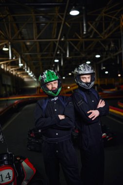 Spor kıyafetleri ve miğferleri olan iki kendine güvenen go kart sürücüsü devrede kollarını kavuşturmuş bekliyor.