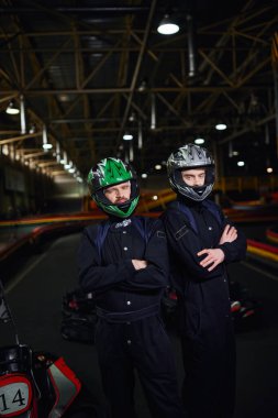 two confident go kart drivers in sportswear and helmets standing with folded arms on circuit clipart