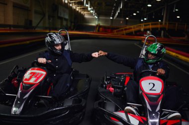 diverse go kart drivers in helmets fist bumping and sitting in sport cars for karting on circuit clipart