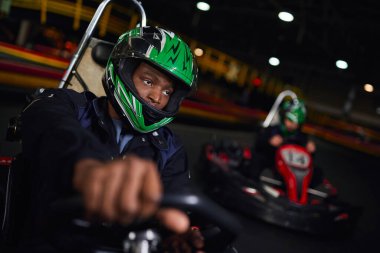 african american man in helmet driving go kart on indoor circuit near friend on blurred backdrop clipart