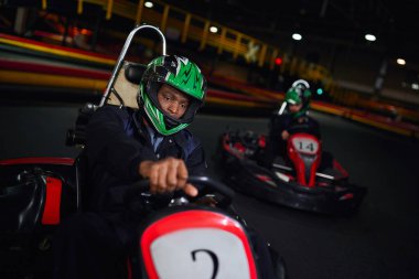 african american driver in helmet driving go kart on circuit near friend on blurred backdrop clipart