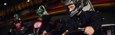 concentrated man driving go kart near diverse drivers in helmets on indoor circuit, banner clipart