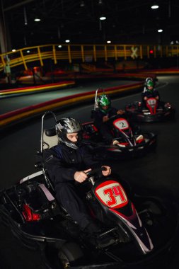 focused man driving go kart near diverse drivers in helmets on indoor circuit, adrenaline and sport clipart