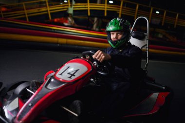 racer in helmet and sportswear driving go kart on indoor circuit, adrenaline and motorsport concept clipart