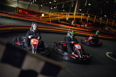 Irklar arası erkekler, puslu ön planda damalı siyah beyaz yarış bayrağının yanında go kart sürüyorlar.